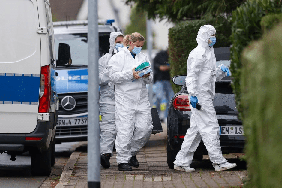 Forensic experts in white hazmat suits and blue face masks and gloves outside a police van and black car.