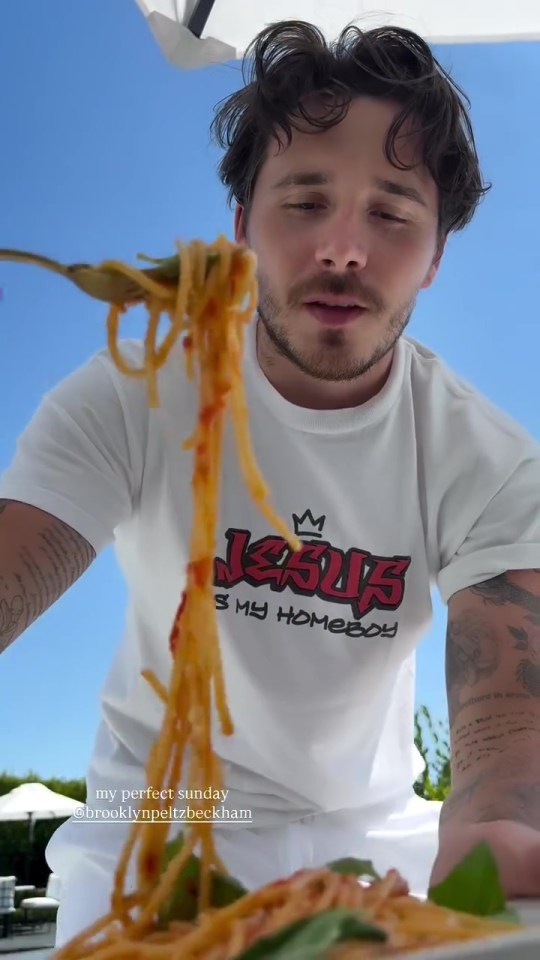 Brooklyn Beckham holding a forkful of spaghetti with tomato sauce, with a clear blue sky in the background.