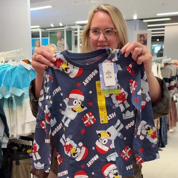 A woman holding up a blue long-sleeved shirt for children with a pattern of cartoon dogs wearing Santa hats and holding presents.