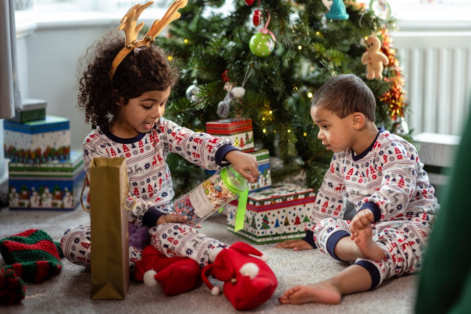 Two young siblings in matching pajamas excitedly opening Christmas gifts on the floor in front of a Christmas tree.