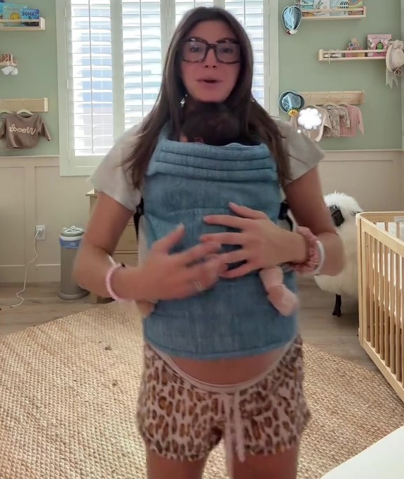 A pregnant woman wearing a baby carrier with an infant, standing in a nursery.
