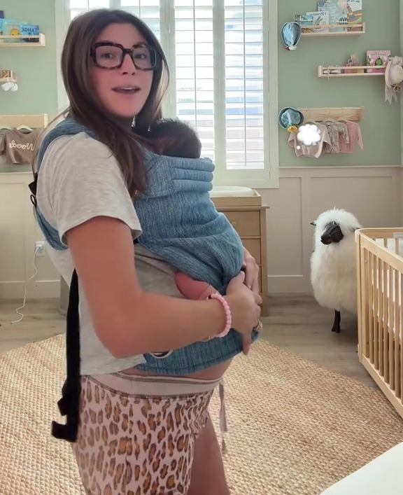 A pregnant woman wearing glasses and cheetah print shorts, carrying a baby in a carrier, standing in a nursery with a crib and a sheep toy.