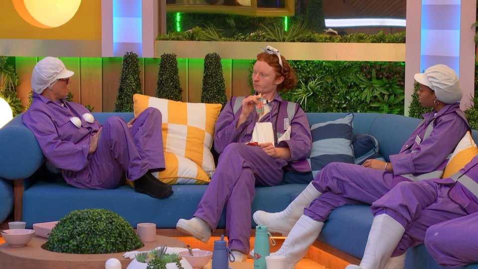 Contestants on Big Brother wearing purple overalls and white hats and boots, sitting on a blue couch.