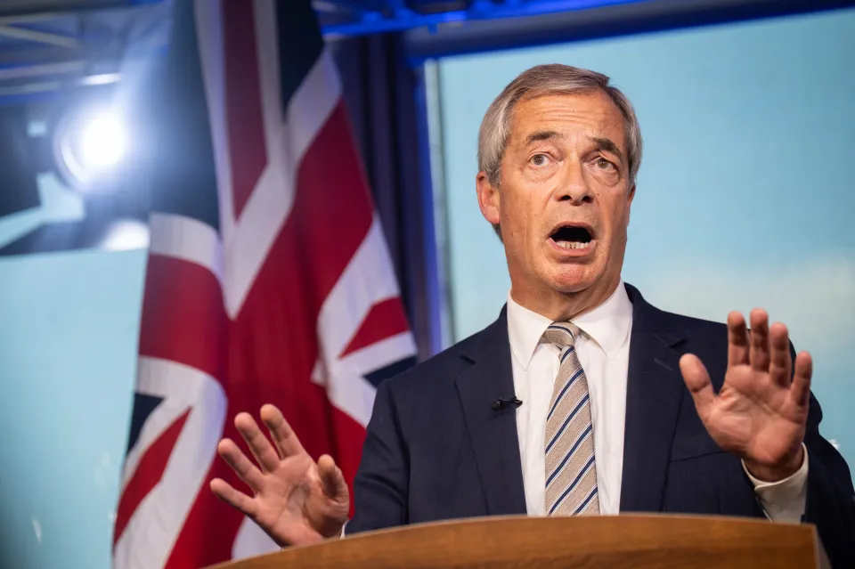 Nigel Farage speaking at a podium with his mouth open and hands raised, with a Union Jack flag behind him.