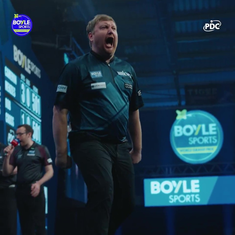 A dart player in a dark shirt and pants yells with excitement on stage, with "Boyle Sports" logos visible in the background.