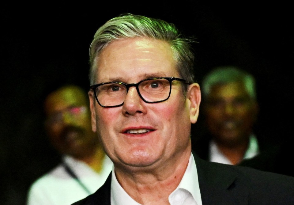 British Prime Minister Keir Starmer wearing glasses, a white shirt, and a dark suit, smiling during his visit to Yash Raj Films in Mumbai, India.