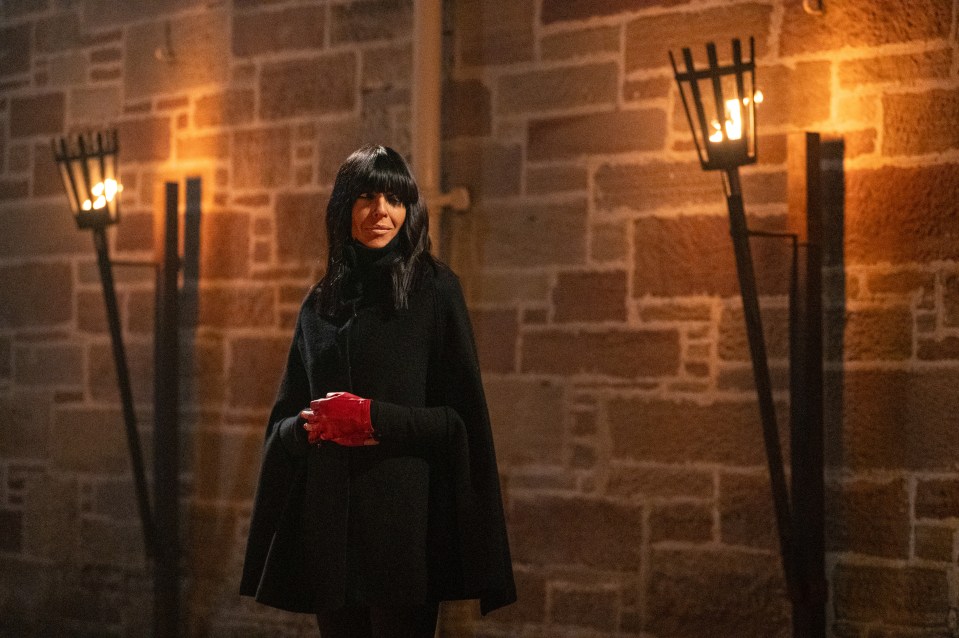 Claudia Winkleman in a black cape and red gloves, standing between two flaming torches against a stone wall.