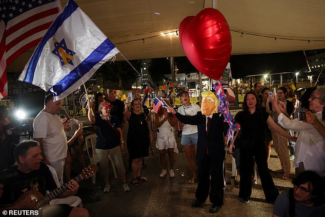 A man in a mask depicting Donald Trump waves an Israeli flag with a yellow ribbon in the center, as people celebrate peace in Tel Aviv
