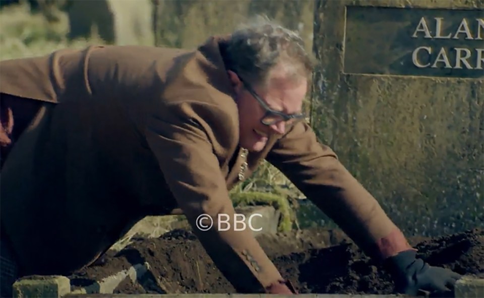 Alan Carr digging in the dirt next to a tombstone with his name on it.