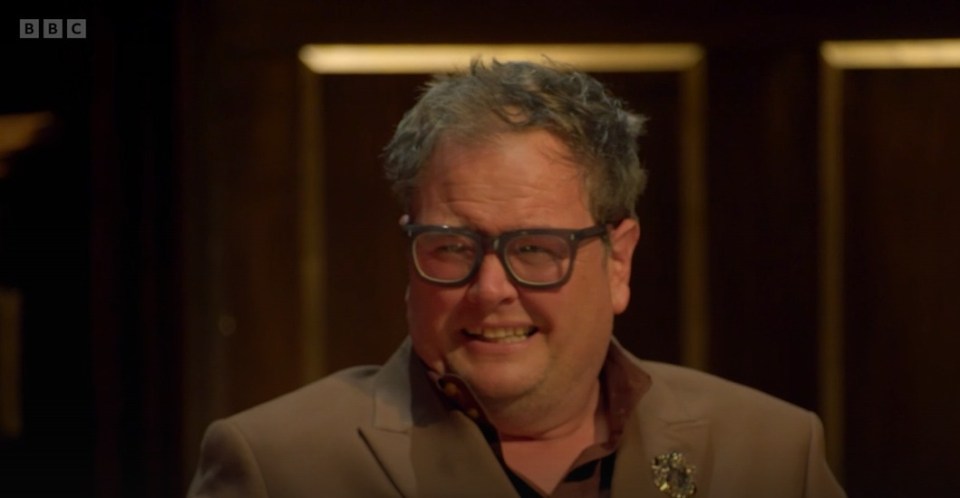 A man with glasses and a gray suit jacket smirks.