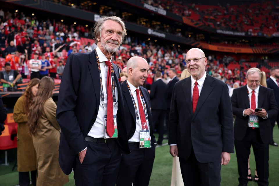 Sir Jim Ratcliffe and Avram Glazer, wearing suits and red ties, looking on at a UEFA Europa League Final in a stadium with spectators in the background.