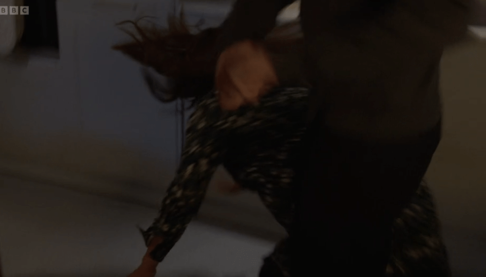 A woman with brown hair falling down, wearing a black and white patterned shirt, is being pushed down by another person.