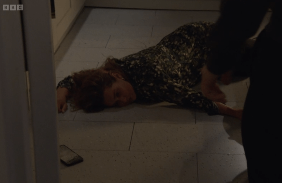 A woman in a printed dress is lying on a light-colored tiled floor, with her head resting on her left arm, and her eyes partially closed. A phone is on the floor near her left side, and a person's leg is visible on the right.