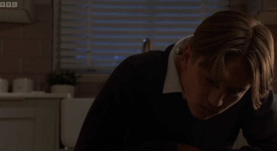 A man with brown hair and a white collared shirt under a black sweater looking down.