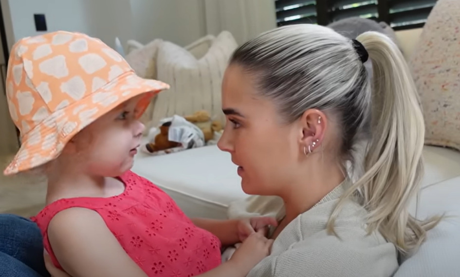 A young girl in a peach-colored hat looking at a woman with blonde hair in a ponytail.