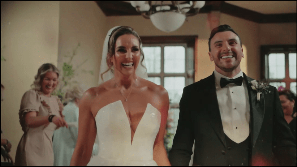 The bride and groom smile as they enter the reception, with the bride’s mother in the background.