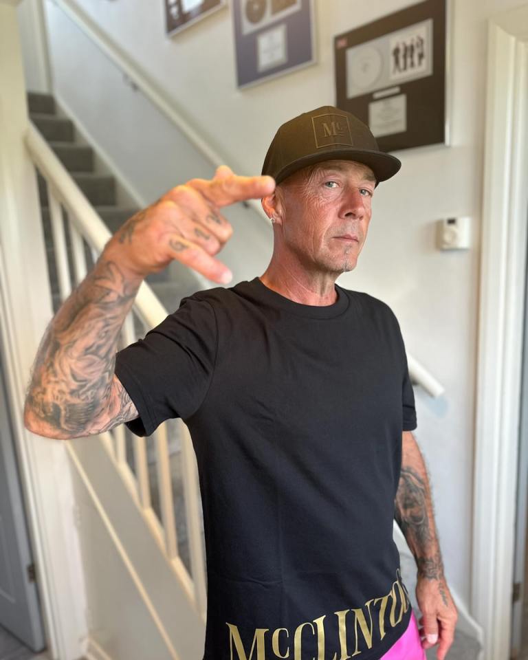 John Hendy of East 17, wearing a black cap and shirt, making a hand gesture.
