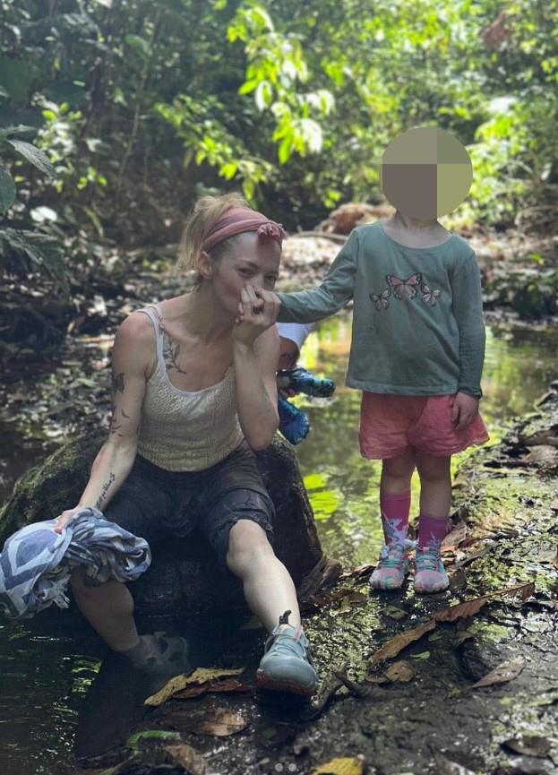 Hannah Spearritt sitting by a stream, with her hand to her mouth, and a child standing beside her.