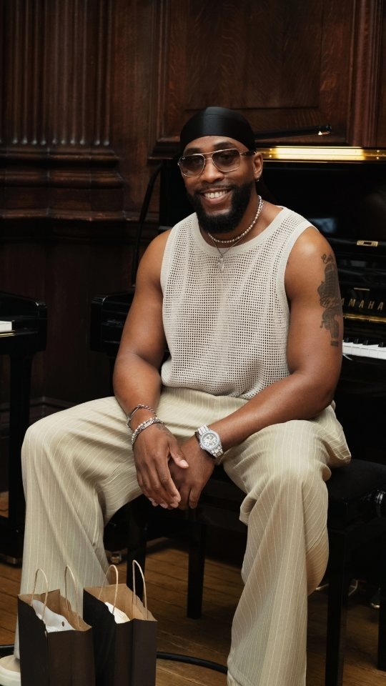 Kule T from MN8 band smiling, wearing a black do-rag, sunglasses, and a light-colored mesh tank top, seated next to a piano.