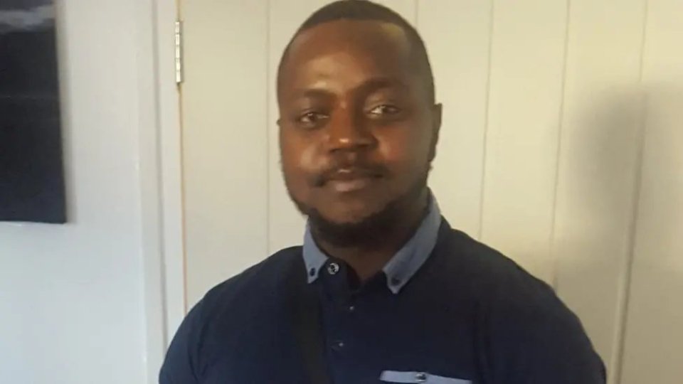 Aron Kato, a black man with short dark hair and a beard, wearing a dark blue collared shirt.