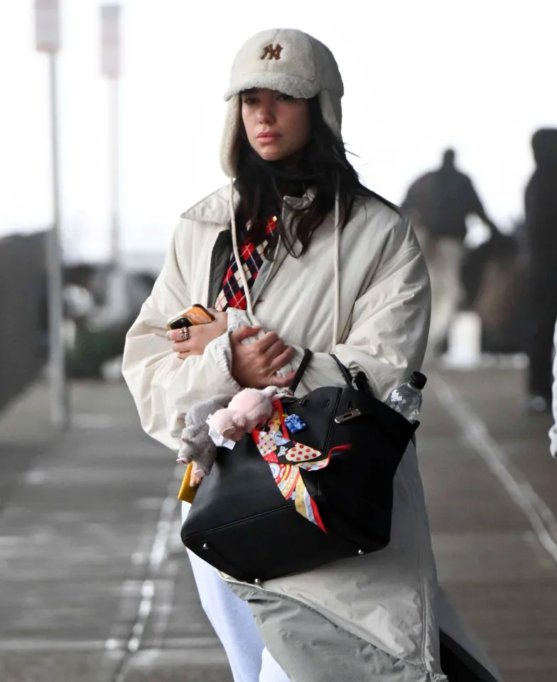Dua Lipa arriving at JFK Airport, wearing a white coat, grey joggers, Ugg boots, and a NY Yankees hat.