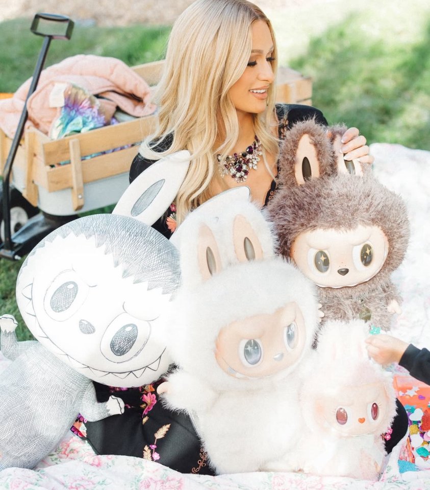 Paris Hilton smiling while holding a brown Labubu toy, surrounded by several other Pop Mart Labubu plush toys.