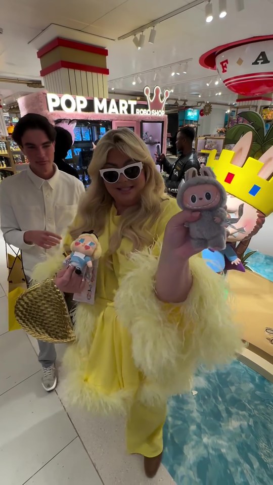 Gemma Collins in a yellow feather-trimmed outfit holding up two collectible dolls in a Pop Mart store.
