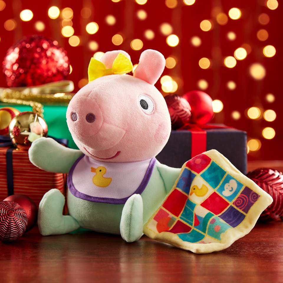 Baby Peppa Pig plush toy with a yellow bow and bib, holding a colorful baby blanket, surrounded by Christmas ornaments and gifts.