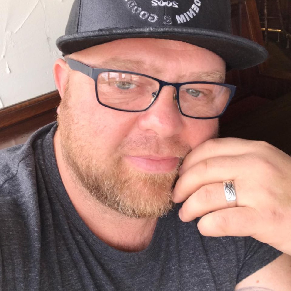 A man with a ginger beard and mustache wearing glasses, a dark gray t-shirt, and a dark gray baseball cap, with a ring on his right ring finger, smiles slightly at the camera.