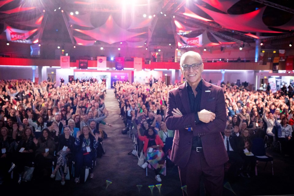 Theo Paphitis standing in front of a large audience, all waving their hands.