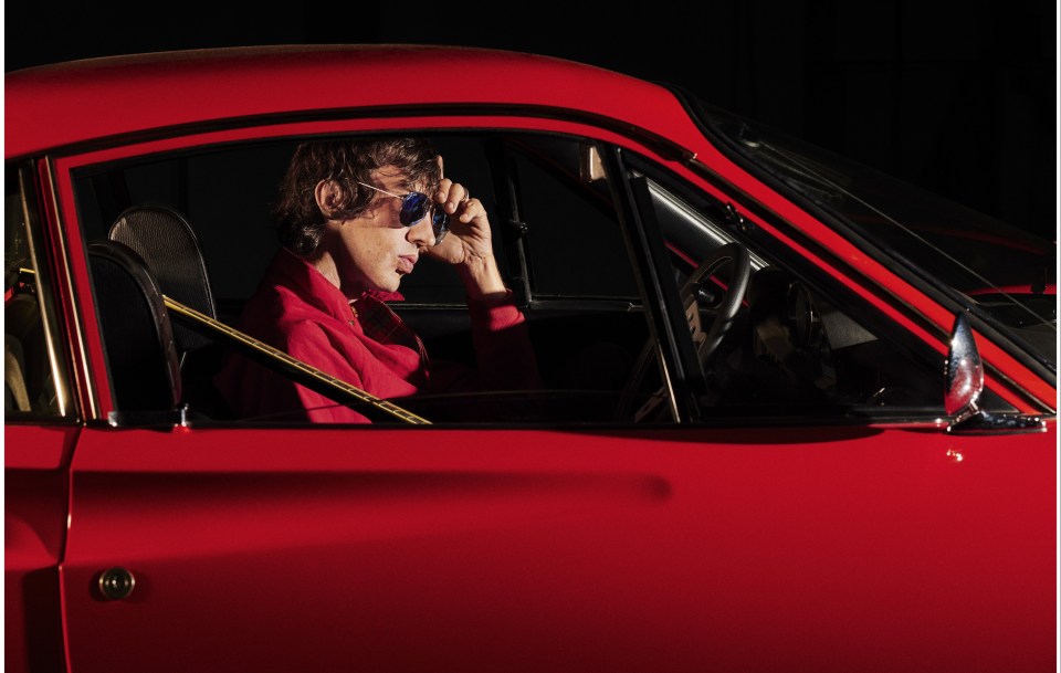 Richard Ashcroft sitting in a red car, adjusting his sunglasses.
