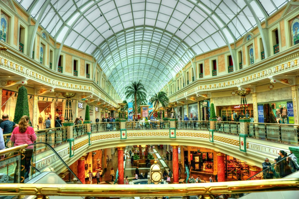 An arched glass roof over a multi-story shopping center with ornate balconies, stores, escalators, people, and palm trees.