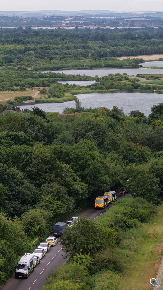 Police cars and vans on a road next to a lake and forest.