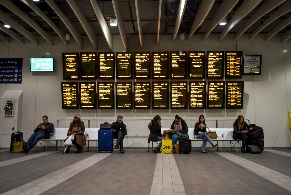 Travelers sit on a bench in front of a departure board at a train station.