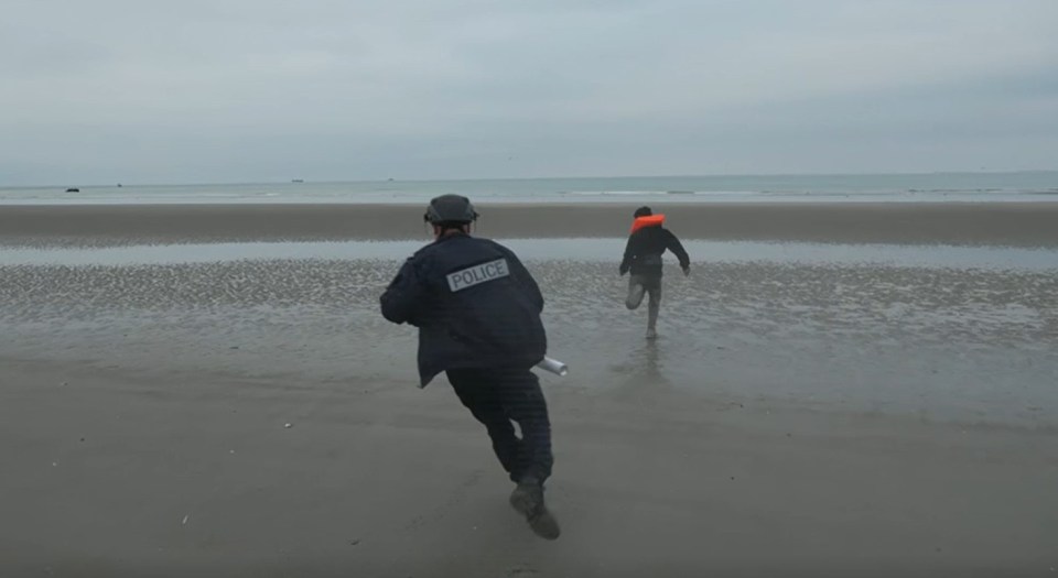 A police officer chasing a migrant wearing a life vest on a sandy beach.