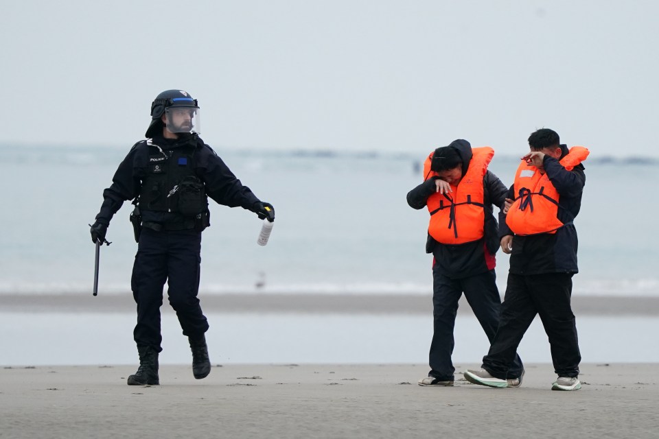 French police officer sprays a substance at two people wearing life jackets on a beach.