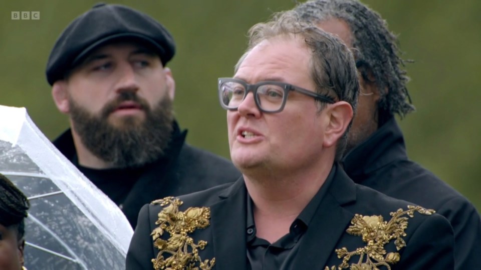 Alan Carr talking while wearing a black blazer with gold embellishments, with other participants in the background.