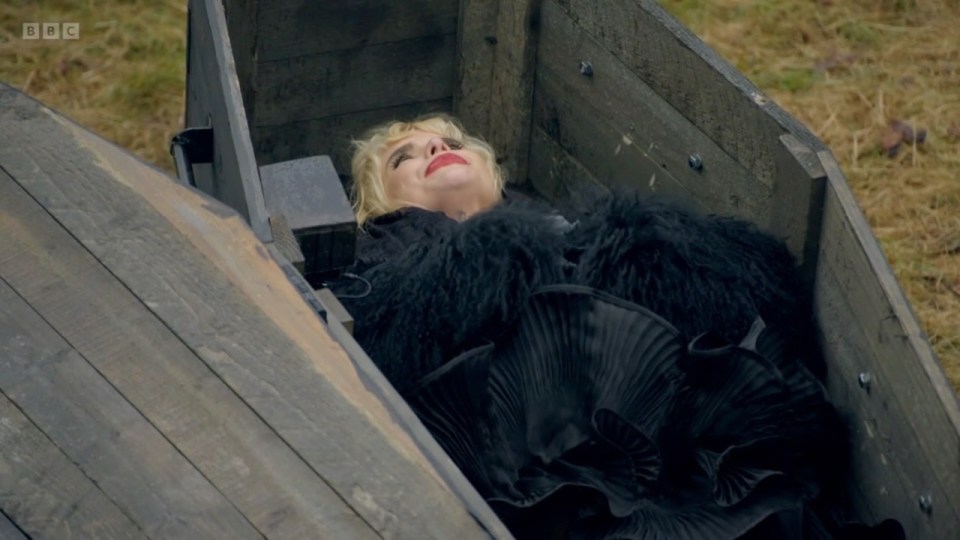 Clare Balding wearing a black coat and red lipstick, lying in a wooden coffin.