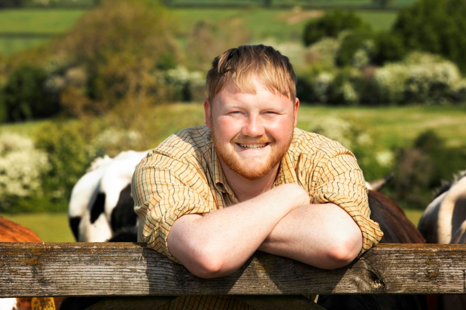Kaleb Cooper in an olive green long-sleeved polo shirt with the initials "KC" embroidered, blue jeans, leaning on a fence post with a field and cloudy sky in the background.