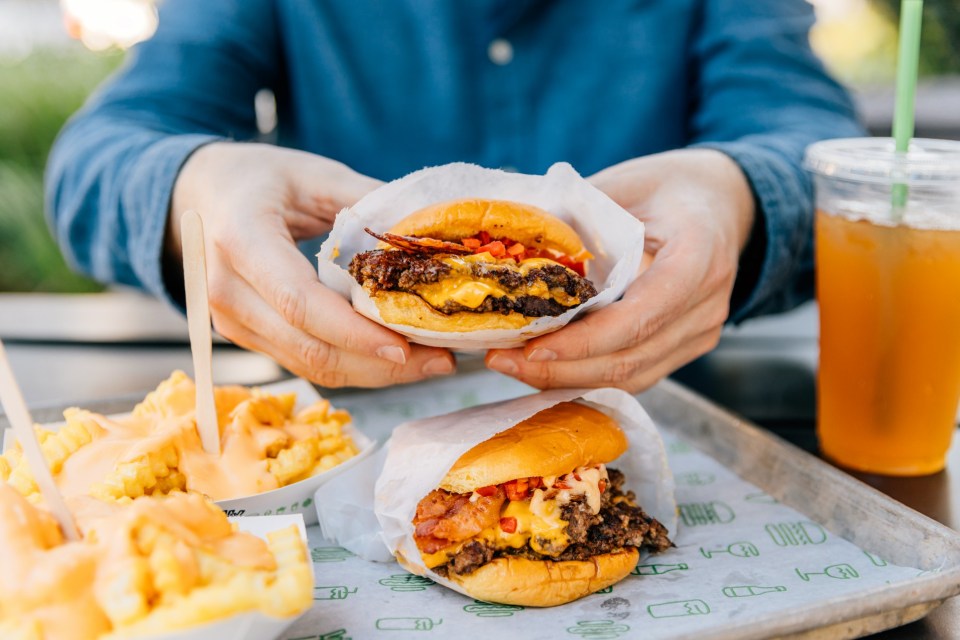 A man holding a cheeseburger, with another cheeseburger, cheese fries, and a drink on a tray.