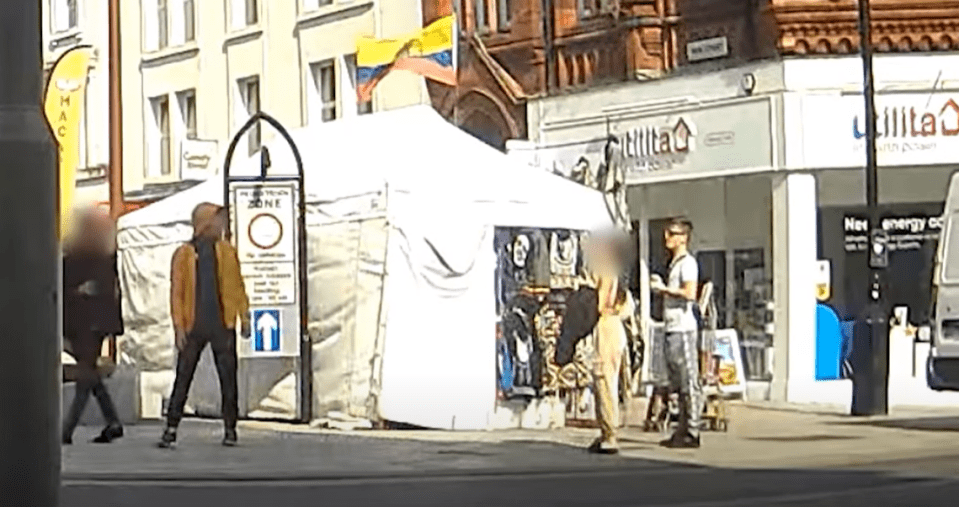 Street scene with a white tent vendor and pedestrians, a yellow, blue, and red flag flying above the tent, and shops in the background.