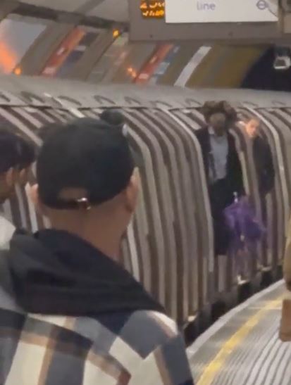 Man in a black baseball cap watching another man and a bicycle on a subway platform.