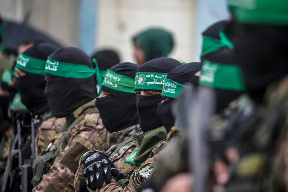 Armed Hamas fighters in camouflage uniforms and black balaclavas, one with a green headband, stand guard.