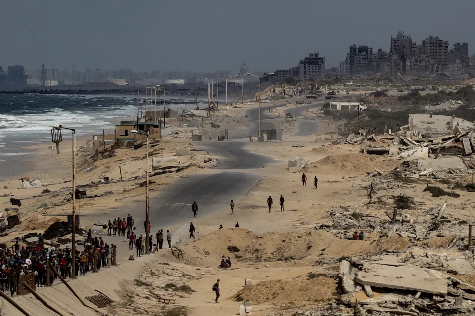 Palestinians turn back on Rashid Street in Deir al-Balah, Gaza, as Israeli forces attack, with the sea on the left and destroyed buildings in the background.