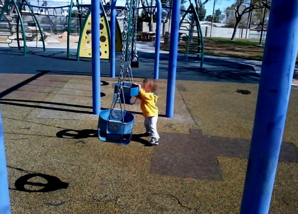 A toddler in a yellow shirt standing next to blue swings on a playground.