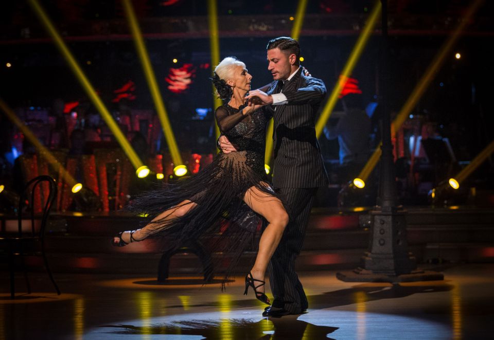 Debbie McGee and Giovanni Pernice dancing in black outfits on a stage lit with yellow spotlights.