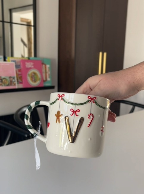 Hand holding a white mug with a gold 'V' and Christmas decorations.