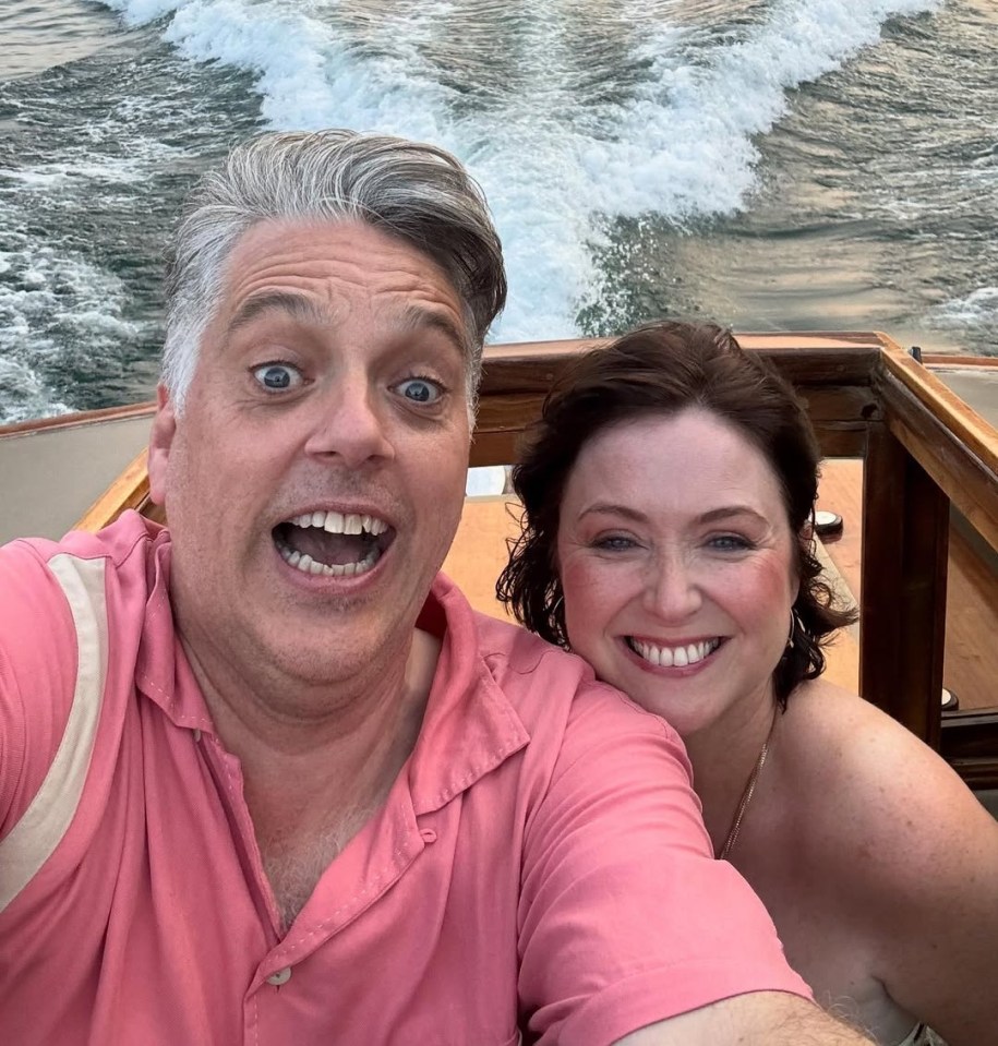 A man in a pink shirt and a woman smile for a selfie on a boat, with a lake and mountains in the background.