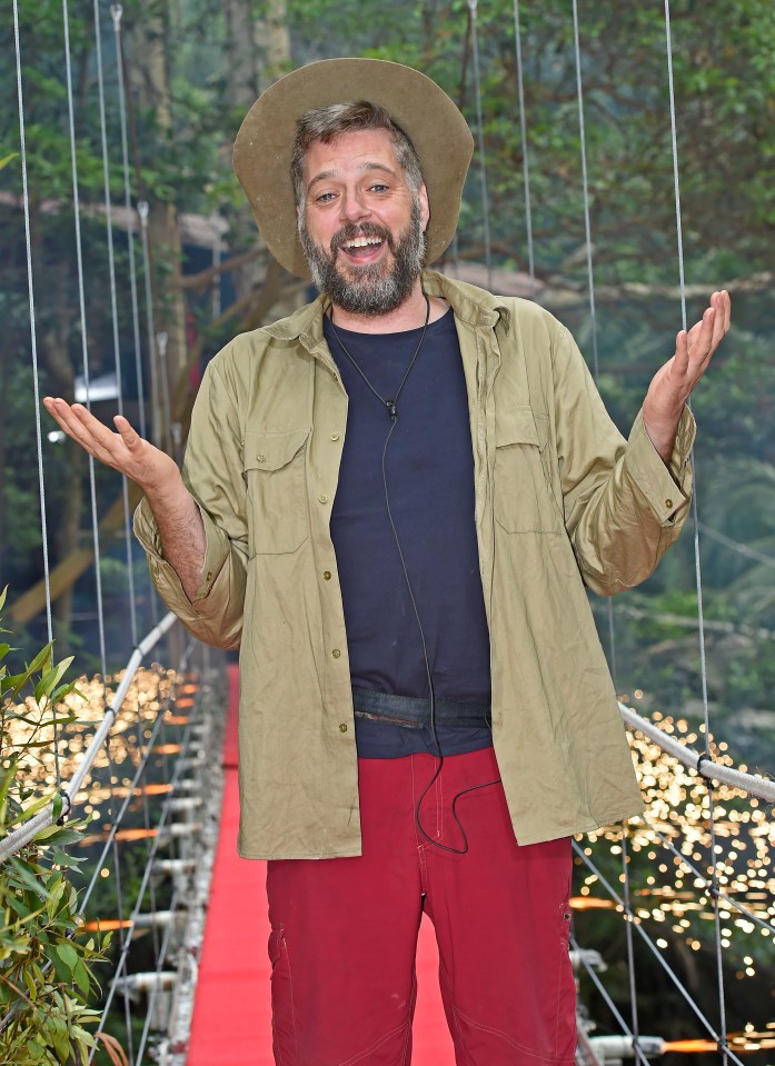 Iain Lee, an evicted contestant from "I'm a Celebrity...Get Me Out of Here!" in 2017, standing on a red carpet bridge with sparkling lights.