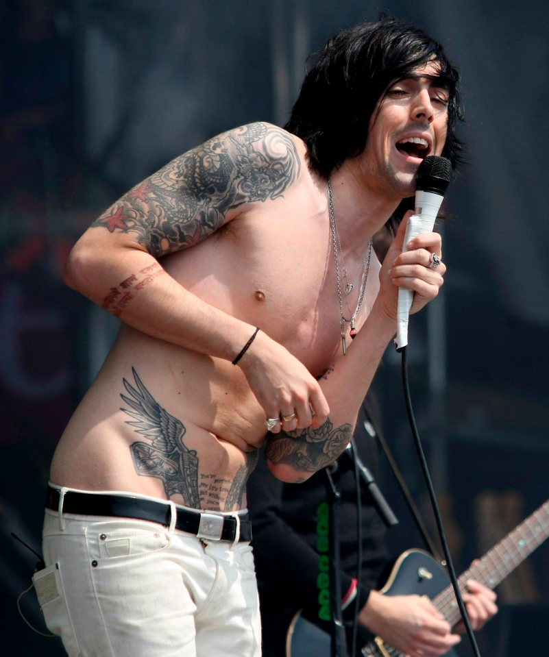 Ian Watkins of Lostprophets singing into a microphone on stage, shirtless with tattoos and white pants.